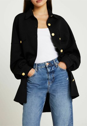 Sort quiltet skjortejakke med guldknapper, der har krave, lange ærmer og en afslappet pasform, båret over en hvid tanktop og blå jeans.