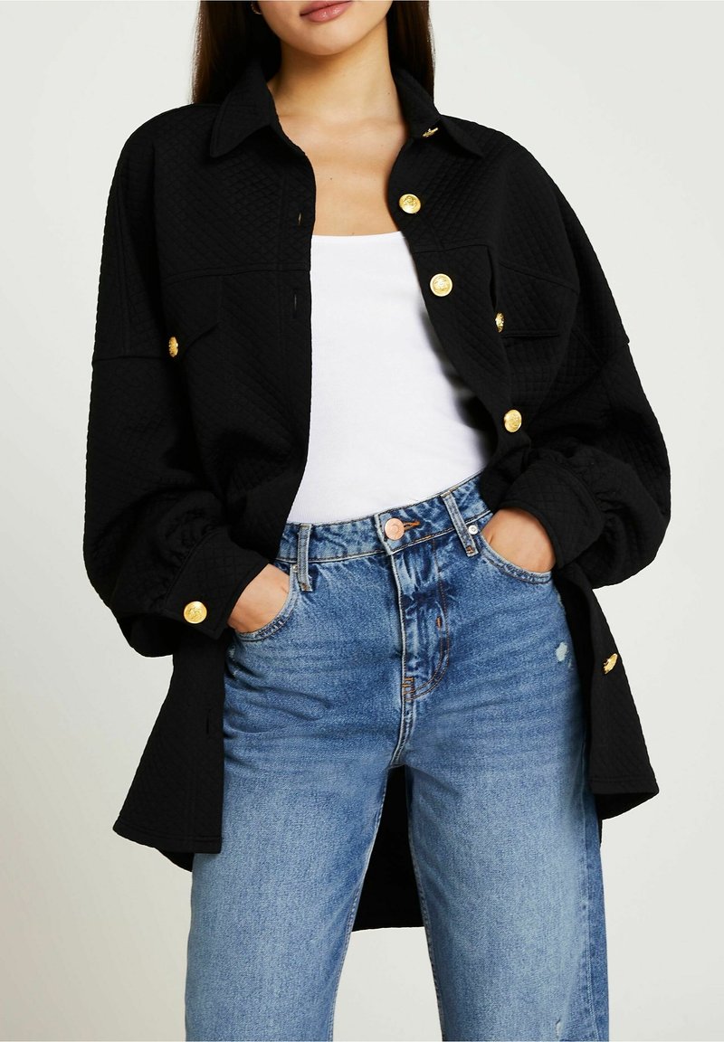 Svart vadderad skjortjacka med guldknappar, med krage, långa ärmar och en avslappnad passform, bärs över en vit linne och blå jeans.