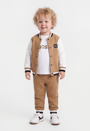 Jeune enfant aux cheveux blonds bouclés portant une veste beige et blanche, un pantalon beige, des baskets blanches et un t-shirt blanc avec le texte "BOSS".