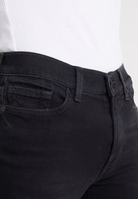 Vaqueros de mezclilla negros Slim Fit, con un bolsillo frontal, trabillas para el cinturón y detalles de costura sutiles. La tela muestra una ligera textura.