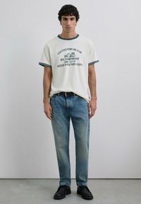 Camiseta blanca con detalles en verde, que presenta una impresión gráfica. Usada con jeans azul claro y zapatos negros con cordones. Fondo liso.