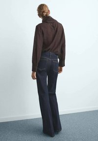 Frau mit blonden Haaren in einem Dutt, die eine dunkelbraune Bluse mit langen Ärmeln und dunkelblaue High-Waist-Jeans trägt, steht vor einer schlichten Wand.
