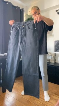 Jeans de mezclilla negra con un diseño de pierna recta, que cuentan con bolsillos frontales, un cierre de botón y un acabado limpio, sostenidos por una persona.