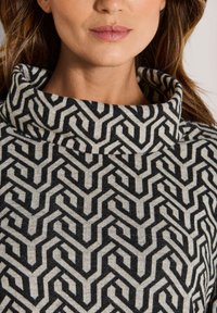 Frau trägt einen schwarz-beigen gemusterten Rollkragenpullover, zeigt das untere Gesicht und den Hals mit lockigem braunem Haar.
