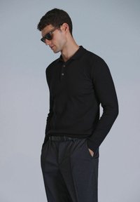 Polo noir à manches longues côtelé avec un col et trois boutons, associé à un pantalon foncé et une ceinture noire. Modèle portant des lunettes de soleil.