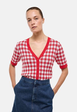 Femme portant un cardigan à manches courtes à carreaux rouges et blancs avec bordure et boutons rouges, associé à une jupe en jean foncé taille haute.