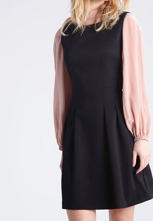 Vrouw draagt een mouwloze zwarte jurk over een lichtroze blouse met lange mouwen, staand tegen een effen lichte achtergrond.