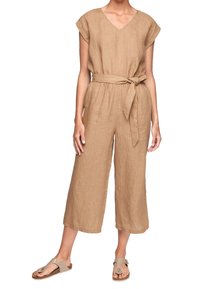 Femme portant une combinaison pantalon beige à jambes larges, manches courtes, col en V, avec une ceinture en tissu nouée à la taille, associée à des sandales beiges.