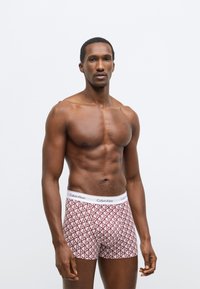 Κόκκινα και λευκά ανδρικά εσώρουχα με γεωμετρικό μοτίβο διαμαντιών, με λευκή ζώνη μέσης που φέρει το λογότυπο "Calvin Klein". Το σώμα είναι μυώδες και καλοσχηματισμένο.