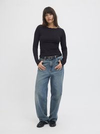 Black long-sleeve top with scalloped neckline; paired with loose light blue denim jeans and black flat shoes, standing against a plain background.