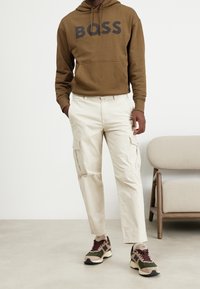 Hnědá mikina s kapucí s logem "BOSS", béžové kalhoty s bočními kapsami a tenisky s maskáčovým vzorem a červenými detaily.