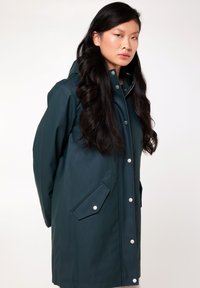 Manteau imperméable vert foncé avec capuche, fermeture à boutons pressions et deux poches avant. Texture lisse et design décontracté.