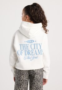 Giovane donna con i capelli ricci raccolti in una coda di cavallo, indossa un hoodie bianco con la scritta "USA La città dei sogni New York" e pantaloni con stampa leopardo, girata di spalle.
