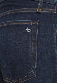 Jeans de denim azul oscuro con un bolsillo trasero que presenta costuras amarillas y un pequeño logotipo bordado en blanco en el lado del bolsillo.