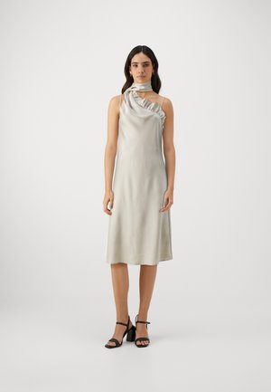 Abito in satin argento asimmetrico con dettagli in ruffle, design senza maniche, lunghezza fino al polpaccio, abbinato a sandali neri con tacco blocco e cinturini.