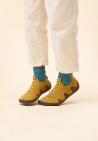 Gelbe gestrickte Schuhe mit braunen Akzenten und einem Reißverschluss, kombiniert mit blauen Socken. Textbranding an der Seite. Weiche, strukturierte Oberfläche.