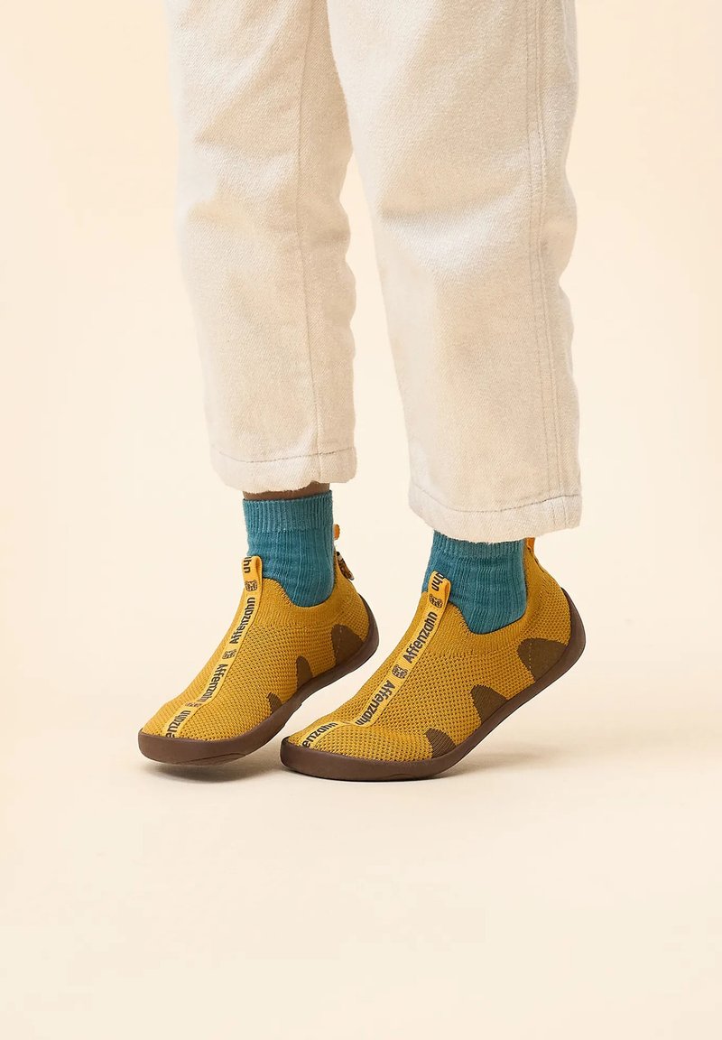 Gelbe gestrickte Schuhe mit braunen Akzenten und einem Reißverschluss, kombiniert mit blauen Socken. Textbranding an der Seite. Weiche, strukturierte Oberfläche.