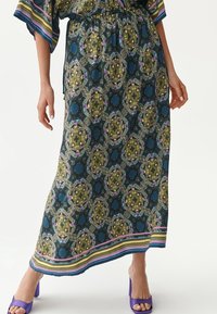 Maxirock mit einem lebhaften, mehrfarbigen Blumen- und geometrischen Muster, einem elastischen Bund und einem gestreiften Saum. Trägt lila Absätze.