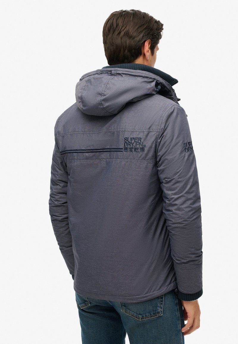 Szara bluza z kapturem o gładkiej fakturze, z zamkiem błyskawicznym, haftowanym logo i ściągaczami na mankietach, w połączeniu z niebieskimi dżinsami.