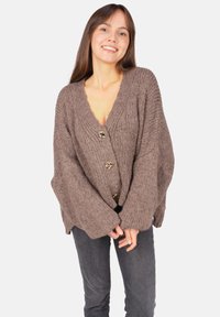 Brauner Strickcardigan mit V-Ausschnitt, vier goldenen Knöpfen und übergroßen Ärmeln. Texturiert und bequem geschnitten. Getragen über grauen Jeans.