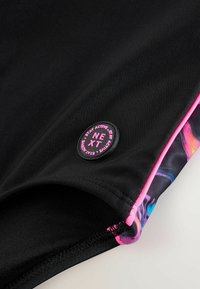 Costumi da bagno neri con una rifinitura rosa. Presentano un'etichetta rotonda nera in tessuto con la scritta "NEXT" in rilievo rosa e un motivo astratto colorato sul lato.