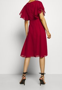 Robe midi rouge avec manches flottantes transparentes, corsage ajusté et jupe évasée. Associée à des sandales à talons aiguilles noires à fines lanières.