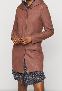 Cappotto di lana marrone, testurizzato, con cappuccio, caratterizzato da una zip frontale, tasche laterali e una vestibilità su misura sopra un abito floreale.