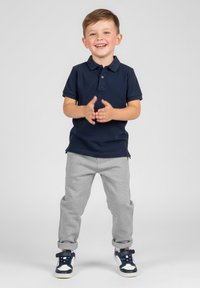 Granatowa koszulka polo, szare spodnie dresowe i granatowe trampki z białymi akcentami. Dziecko stoi z rękami złożonymi razem na neutralnym tle.