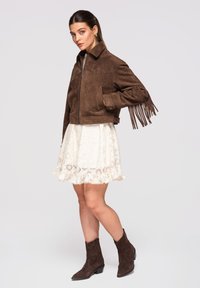 Donna che indossa una giacca con frange in camoscio marrone, una gonna di pizzo bianca e stivaletti in camoscio marrone abbinati, in piedi davanti a uno sfondo semplice.