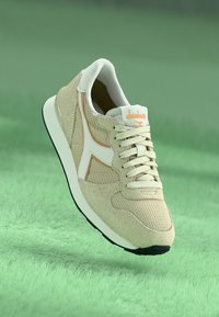 Beige athletic shoe with mesh and suede materials, white accents, laces, and a black outsole. Features a subtle logo on the tongue.