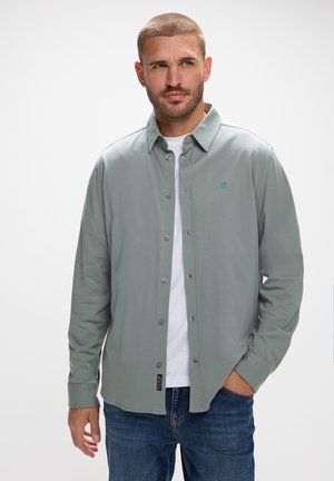 Jasnozielona koszula z długim rękawem, z zapinanym przodem, kołnierzykiem i subtelnym logo. Nosi się ją na białym T-shircie, w połączeniu z niebieskimi dżinsami.
