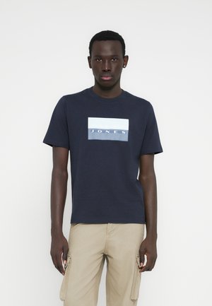 Marineblauw katoenen T-shirt met een rechthoekige witte en blauwe afbeelding waarop "JONES" staat, horizontaal over de borst. Korte mouwen en een ronde hals.