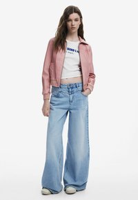 Rosa cropped Bomberjacke über einem weißen T-Shirt mit blauer Schrift, kombiniert mit hellblauen, weiten Jeans und beigen Sneakers.