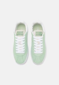 Žalios zomšinės sportbačiai su baltais raišteliais, minimalistinio dizaino, pasižymintys kreminės spalvos liežuviu su logotipo detalėmis ir balta guma padanga.