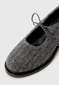 Close-up of a gray knit fabric shoe with a rounded toe, black sole, and a tied gray lace bow on a plain background.