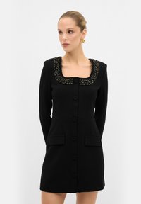 Robe noire avec un décolleté carré, ornée d'un col perlé, dotée de manches longues, de boutons sur le devant et de poches latérales.