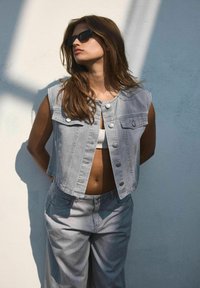 Traper prsluk svijetlosive boje s okruglim izrezom, prednjim džepovima i kopčanjem na dugmad, u kombinaciji s širokim sivim hlačama. Glatka tekstura.