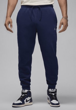 Mężczyzna w granatowych spodniach dresowych z troczkiem i czarno-białych butach Nike Air, stojący na szarym tle.