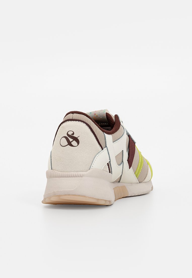 Baskets de sport avec un dessus synthétique beige, des accents marron, des rayures jaunes et une semelle texturée avec un logo au talon.