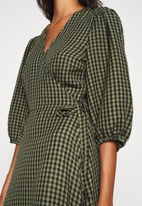 Robe portefeuille à carreaux verts et noirs avec manches bouffantes, présentant une taille cintrée et un tissu texturé dans un design léger.