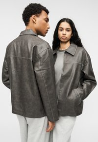 Γκρι τζάκετ από ψεύτικο δέρμα με γιακά, φαρδιά γραμμή, εμφανείς ραφές και πλαϊνές τσέπες. Απλός σχεδιασμός, ματ υφή.