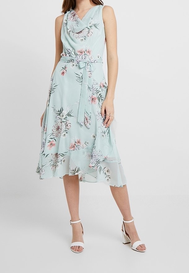 Maxi-jurk van licht mintgroene stof met bloemenpatronen. Cowled halsontwerp, mouwloos, met een ceintuur in de taille en een gelaagde zoom. Gecombineerd met witte hakken.