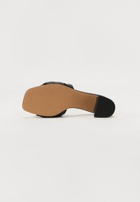 Black heeled shoe with a square toe and a textured black strap. The sole is light brown with a smooth, slightly ridged surface.