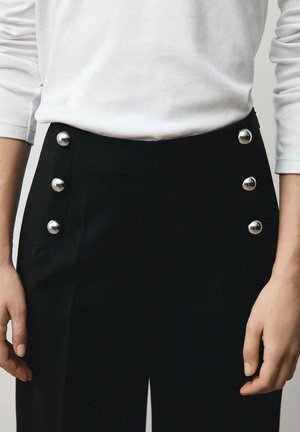 Pantalon noir avec six boutons en argent brillants sur le côté gauche. Fabriqué en tissu lisse avec une coupe ajustée, assorti à une chemise blanche à manches longues.