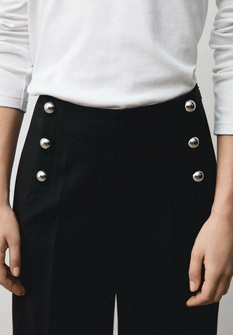 Pantalon noir avec six boutons en argent brillants sur le côté gauche. Fabriqué en tissu lisse avec une coupe ajustée, assorti à une chemise blanche à manches longues.