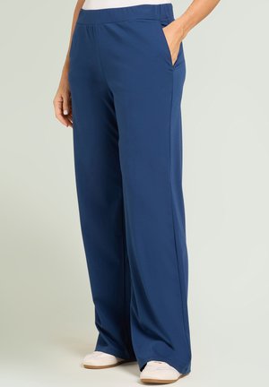 Femme portant un pantalon bleu marine ample avec poches latérales et des baskets blanches, main dans la poche, debout sur un fond clair.