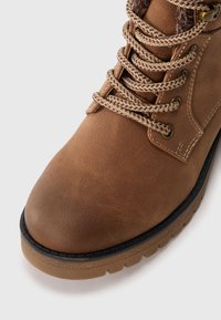 Bottes en cuir marron avec une finition texturée, dotées de lacets beige et d'œillets en métal. La semelle est en caoutchouc avec un motif de semelle pour une meilleure adhérence.