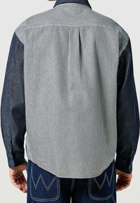 Pruhovaná košile s modrým denimovým yoke a dlouhými rukávy. Má vertikální bílou a černou pruhy, zapínání na knoflíky a sklady na zádech.