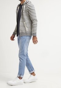 Hoodie a righe in crema e blu navy, materiale leggero. Abbinato a jeans azzurri chiari e sneakers bianche con dettagli testurizzati.