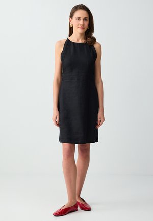 Donna in piedi che indossa un vestito nero senza maniche fino al ginocchio e ballerine rosse su uno sfondo grigio chiaro uniforme.